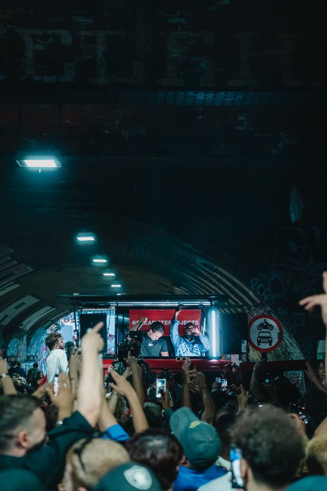 The lit DJ booth on the G-Class at the far end of a brick railway tunnel, hands raised in the foreground