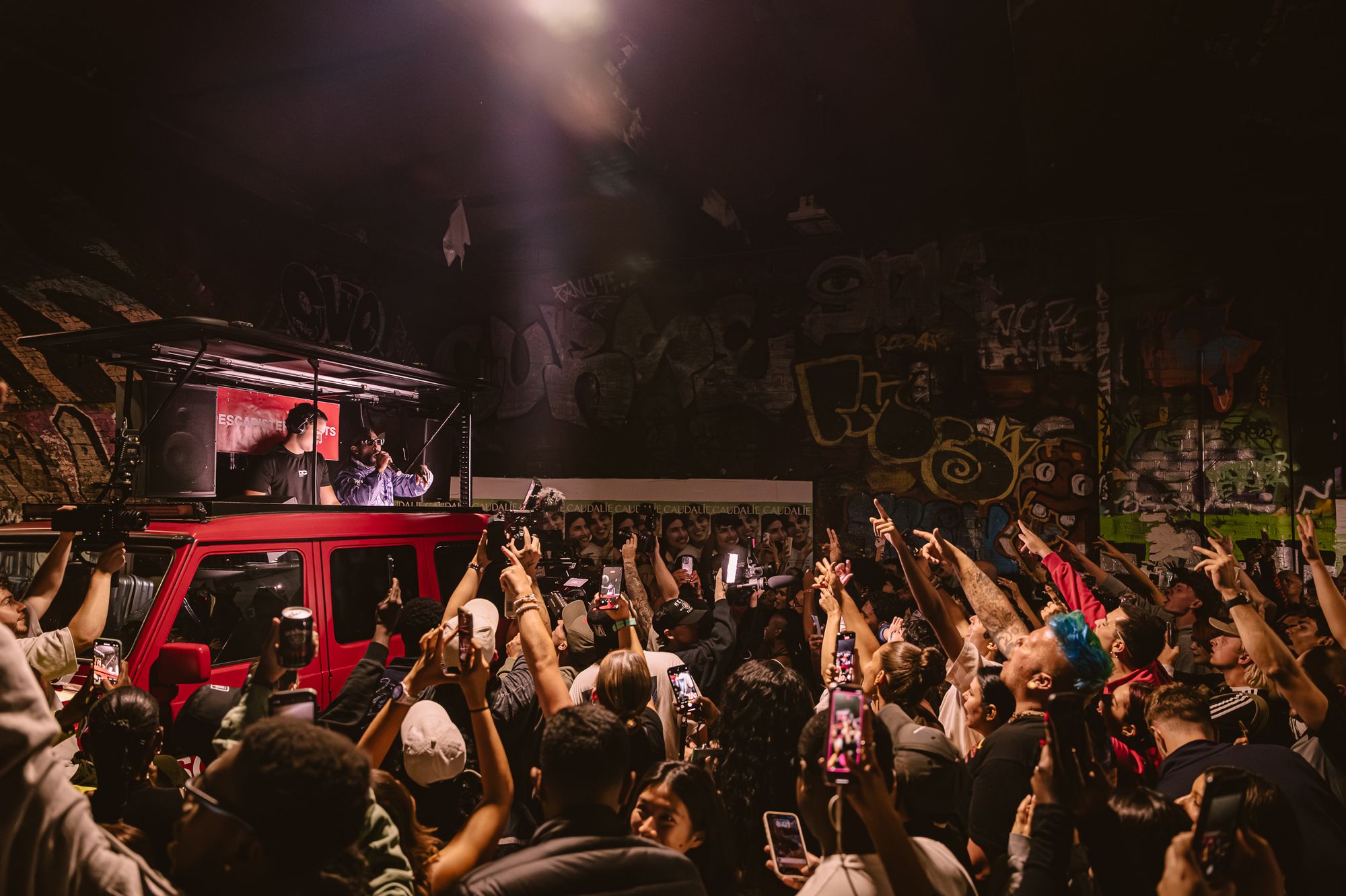 A red G-Class with a DJ booth on its roof surrounded by a crowd in a graffiti tunnel