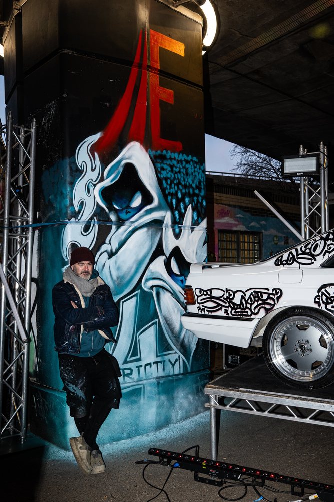 Stockeartworks beside his hooded-figure mural on a Westway column, next to the white art car on its stage deck