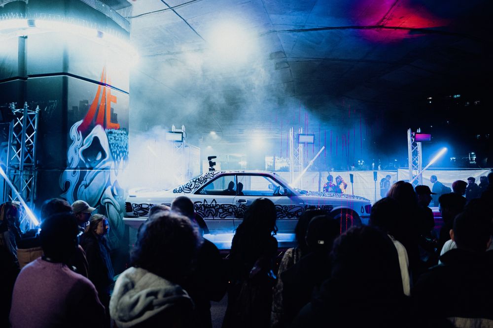 A white car covered in black line drawings, lit blue through haze, with the crowd in front