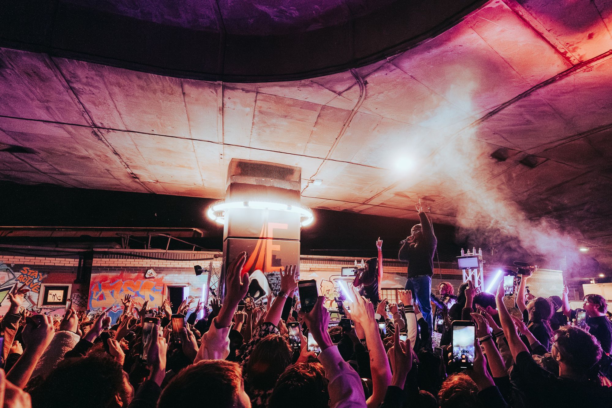 Hands and phones raised towards two performers beside a lit column under the flyover